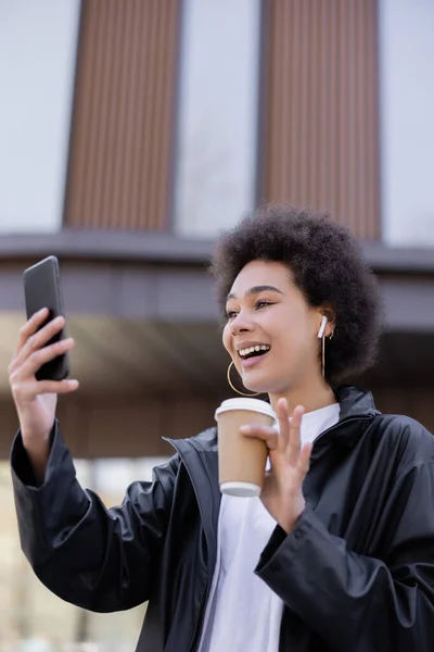 Joyeuse femme afro-américaine dans un écouteur tenant une tasse en papier et ayant un chat vidéo sur smartphone à l'extérieur — Photo de stock