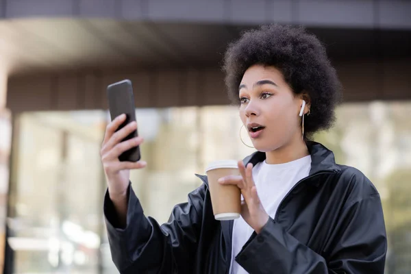 Femme afro-américaine choquée dans un écouteur tenant une tasse en papier et ayant un chat vidéo sur smartphone à l'extérieur — Photo de stock