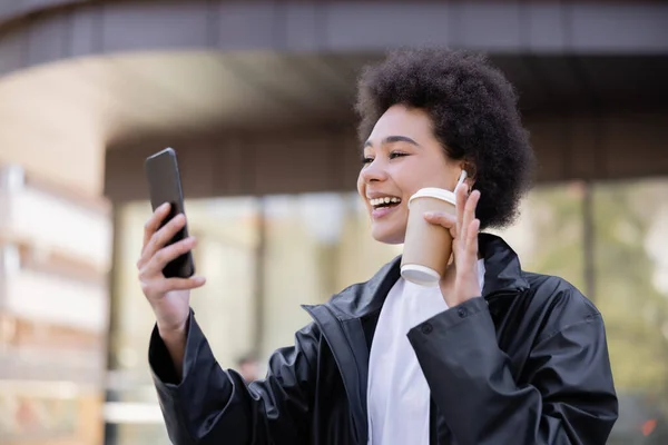 Cheerful african american woman in earphone holding paper cup and having video chat on smartphone — Photo de stock