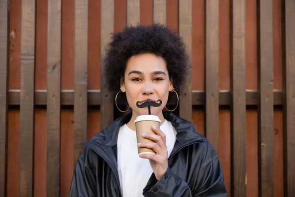 Curly african american woman holding paper cup and straw with moustache near fence — Stock Photo