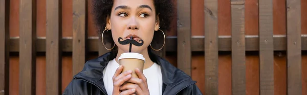 Curly african american woman drinking takeaway beverage from straw with moustache near fence, banner — Stock Photo