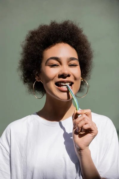 Sorridente donna afroamericana mangiare paglia gelatina vicino al muro verde — Foto stock