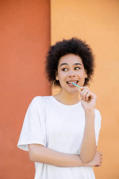 Joyful african american woman eating tasty jelly straw near orange wall - foto de stock