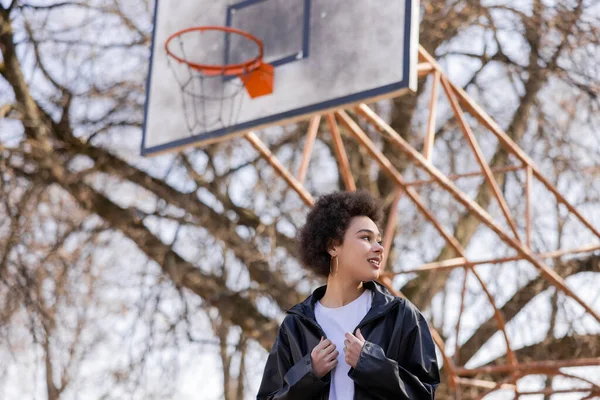 Vue à angle bas de femme afro-américaine heureuse en veste debout sur le terrain de basket-ball extérieur — Photo de stock