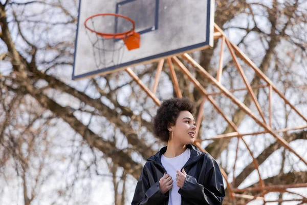 Low angle view of happy african american woman in jacket standing on basketball outdoor court — стоковое фото