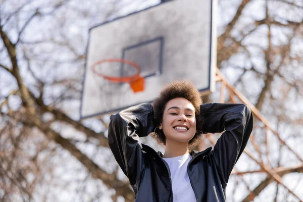 Low angle view of cheerful african american woman in jacket standing on basketball outdoor court — Stockfoto