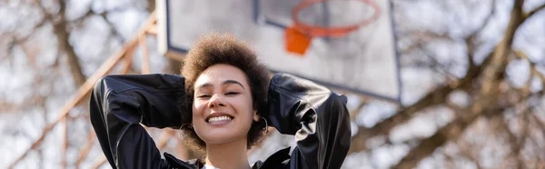 Низький кут зору щасливої афроамериканки в куртці, що стоїть на баскетбольному майданчику, банер — стокове фото