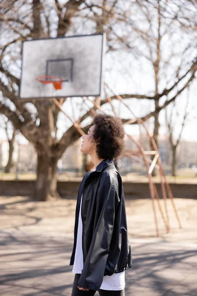 Vue latérale de femme afro-américaine heureuse en veste près du terrain de basket-ball en plein air — Photo de stock