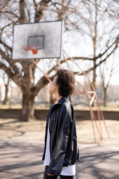 Side view of happy african american woman in jacket near basketball outdoor court — стоковое фото