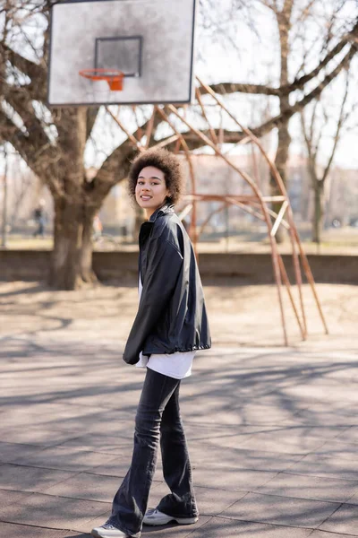 Vue pleine longueur de femme afro-américaine heureuse en veste près du terrain de basket-ball extérieur — Photo de stock