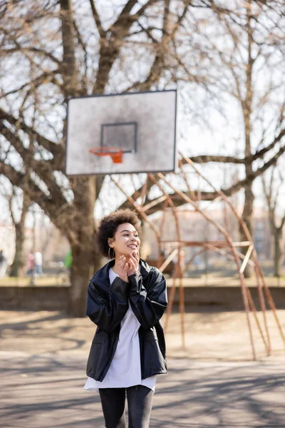 Joyful african american woman in jacket near basketball outdoor court — стоковое фото