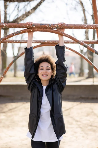 Joyful african american woman in jacket hanging on horizontal bar in rusty outdoor gym — стоковое фото