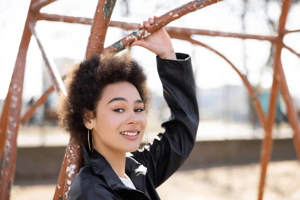 Pleased african american woman in jacket smiling at rusty outdoor gym — Stock Photo