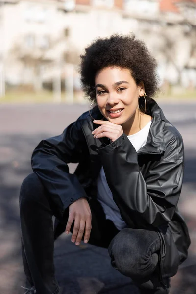 Fröhliche afrikanisch-amerikanische Frau in Jacke sitzt auf Asphalt — Stockfoto