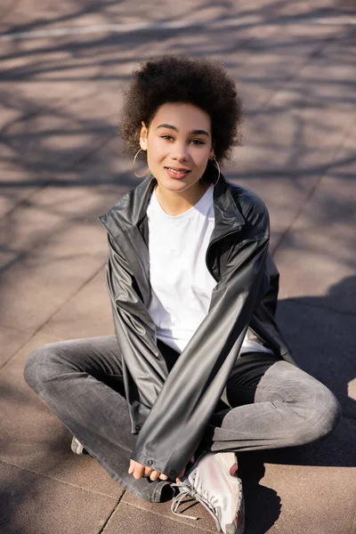 Positive african american woman in jacket sitting on asphalt — Stock Photo