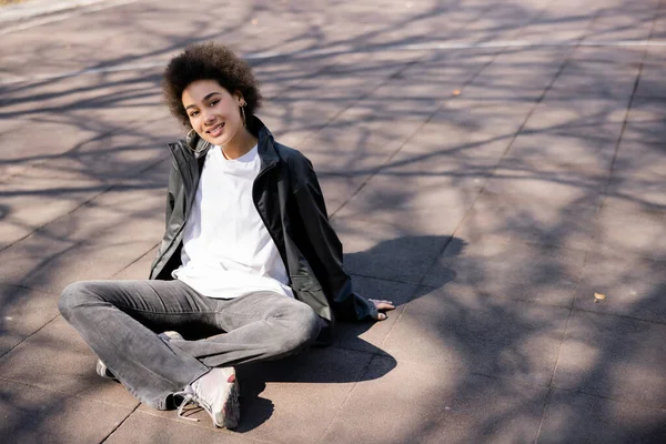 Pleine longueur de souriante femme afro-américaine en veste assis avec les jambes croisées sur l'asphalte avec des ombres — Photo de stock