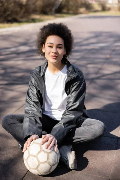 Full length of curly african american woman in jacket sitting with crossed legs and ball on asphalt — Stock Photo