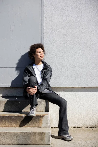 Happy african american woman in jacket sitting on stairs near wall — Stock Photo