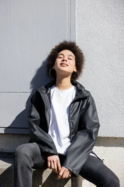 Pleased african american woman in jacket sitting on stairs near wall — стоковое фото