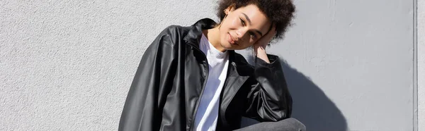 Mujer afroamericana feliz en chaqueta sonriendo cerca de la pared, pancarta - foto de stock