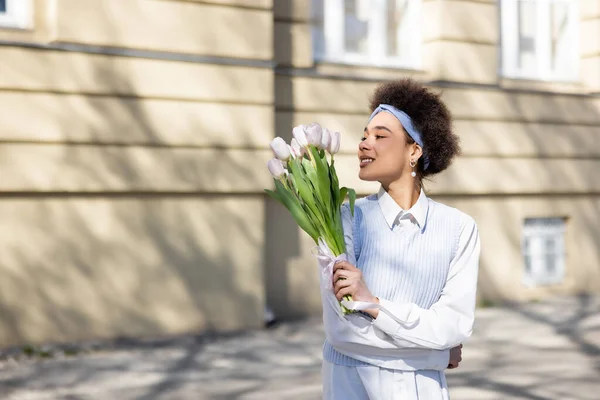 Joyful african american woman looking at bouquet of tulips on street — Stock Photo