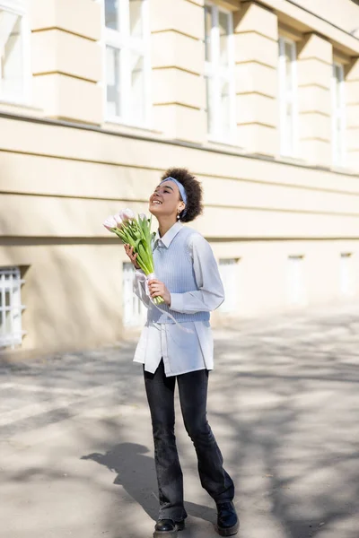 Cheerful african american woman holding bouquet of tulips and walking on street — Stock Photo