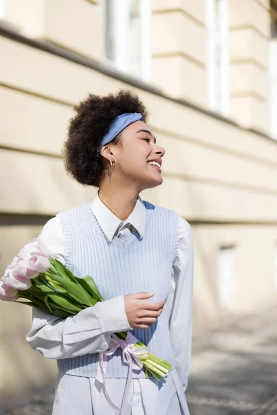 Positive young african american woman in blue vest and white shirt holding bouquet of tulips — Stockfoto