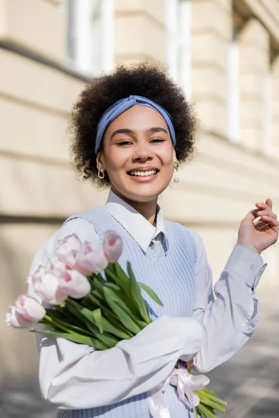 Cheerful young african american woman in blue vest and white shirt holding bouquet of tulips - foto de stock