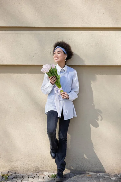Longitud completa de alegre joven afroamericana mujer en chaleco azul y camisa blanca celebración ramo de tulipanes - foto de stock