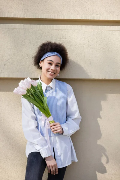 Alegre mujer afroamericana en chaleco azul y camisa blanca sosteniendo ramo de tulipanes cerca de la pared - foto de stock