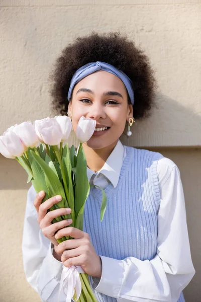 Sonriente mujer afroamericana en chaleco azul y camisa blanca sosteniendo ramo de tulipanes cerca de la pared - foto de stock