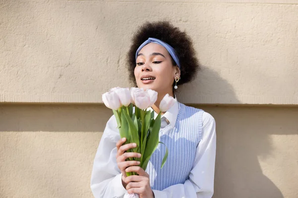 Mujer afroamericana asombrada en chaleco azul y camisa blanca sosteniendo ramo de tulipanes cerca de la pared - foto de stock