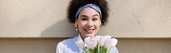 Joyful african american woman in blue vest and white shirt holding bouquet of tulips near wall, banner — Stock Photo