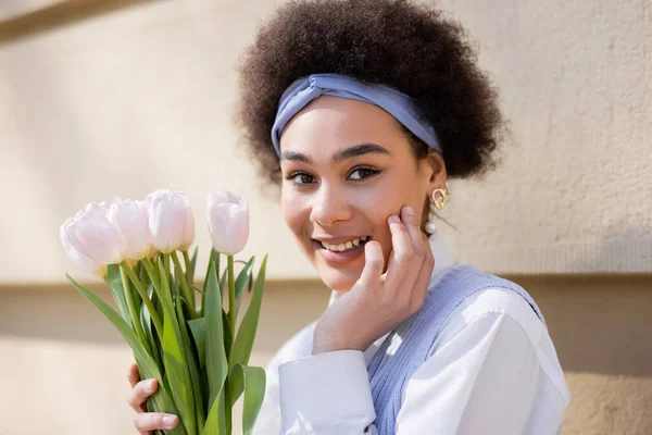 Smiling african american woman in blue headband holding bouquet of tulips near wall — Stock Photo