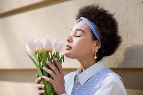 Lockige afrikanisch-amerikanische Frau in blauem Stirnband duftenden Tulpen in der Nähe der Wand — Stockfoto