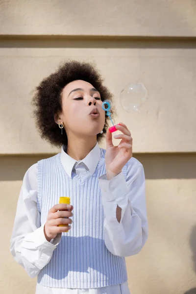 Curly african american woman in blue vest and white shirt blowing soap bubble - foto de stock