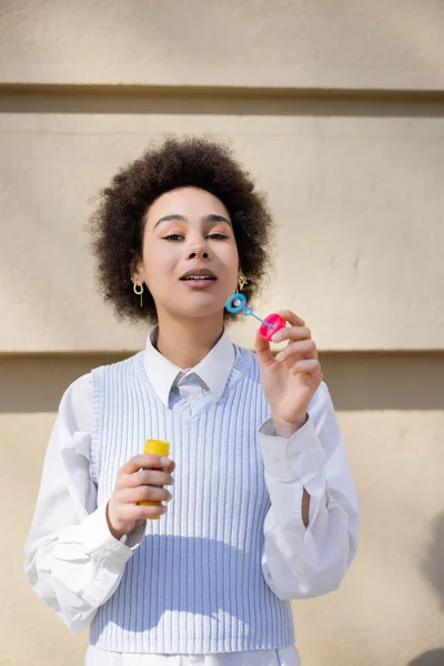 Curly african american woman in blue vest and white shirt holding bubble wand — Stockfoto