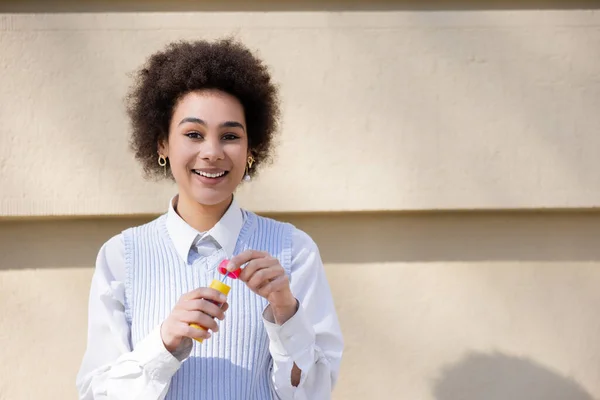Happy african american woman in blue vest and white shirt holding bottle with soap bubbles — Foto stock