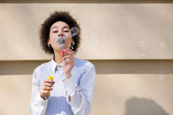 Curly african american woman holding bottle and blowing soap bubbles near wall — Stock Photo