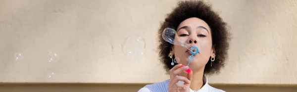 Curly african american woman blowing soap bubble near wall, banner — Stock Photo
