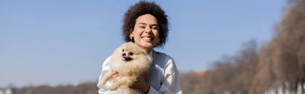 Mujer afroamericana feliz sosteniendo en brazos perro pomerania, bandera - foto de stock