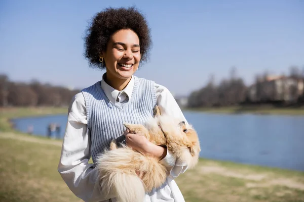 Happy african american woman with closed eyes holding pomeranian spitz — Stock Photo