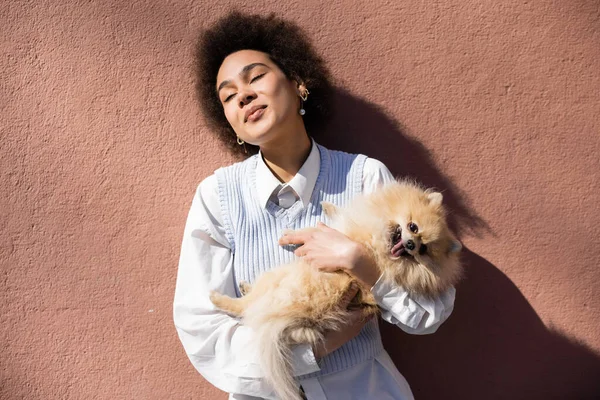 Cheerful african american woman in blue vest holding in arms pomeranian spitz near wall — Stock Photo