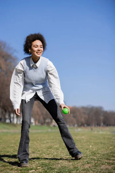 Piena lunghezza di donna afro-americana sorridente in camicia e gilet che tiene la palla nel parco — Foto stock