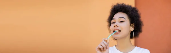 Frisée et jeune femme afro-américaine manger bâton de gelée près du mur orange, bannière — Photo de stock