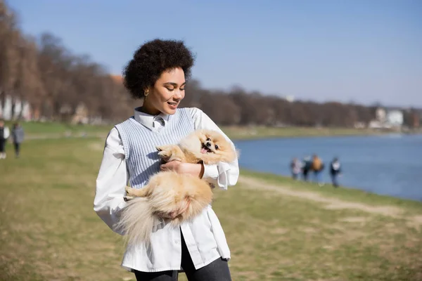 Joyful african american woman in stylish outfit holding pomeranian spitz in park — Photo de stock