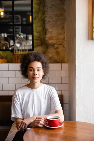 Mujer afroamericana en pendientes de aro mirando a la cámara en la cafetería - foto de stock