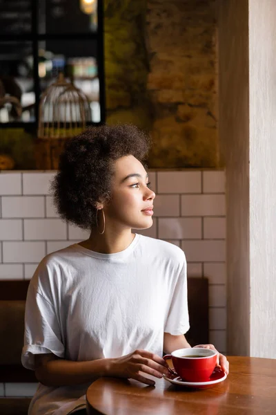 African american woman in hoop earrings and white t-shirt holding cup of coffee — стокове фото