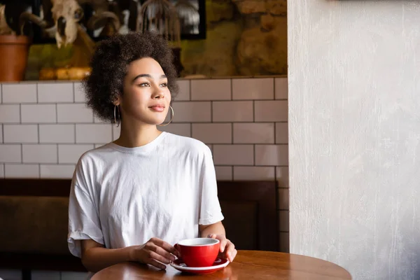 Dreamy african american woman in hoop earrings and white t-shirt holding cup — Stock Photo