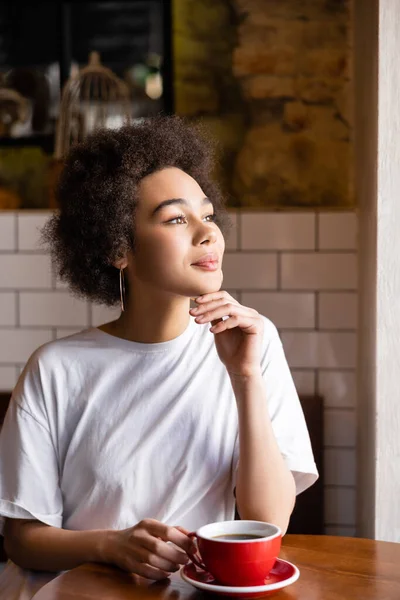 Pensive african american woman in white t-shirt holding cup while looking away - foto de stock